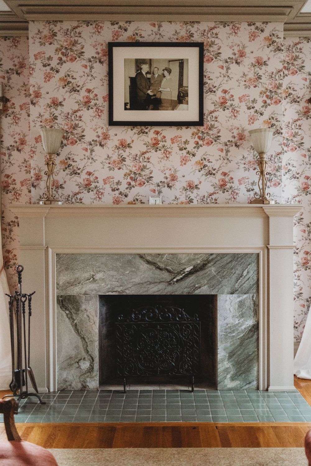A marble fireplace with floral wallpaper and vintage frame above in an elegant room.