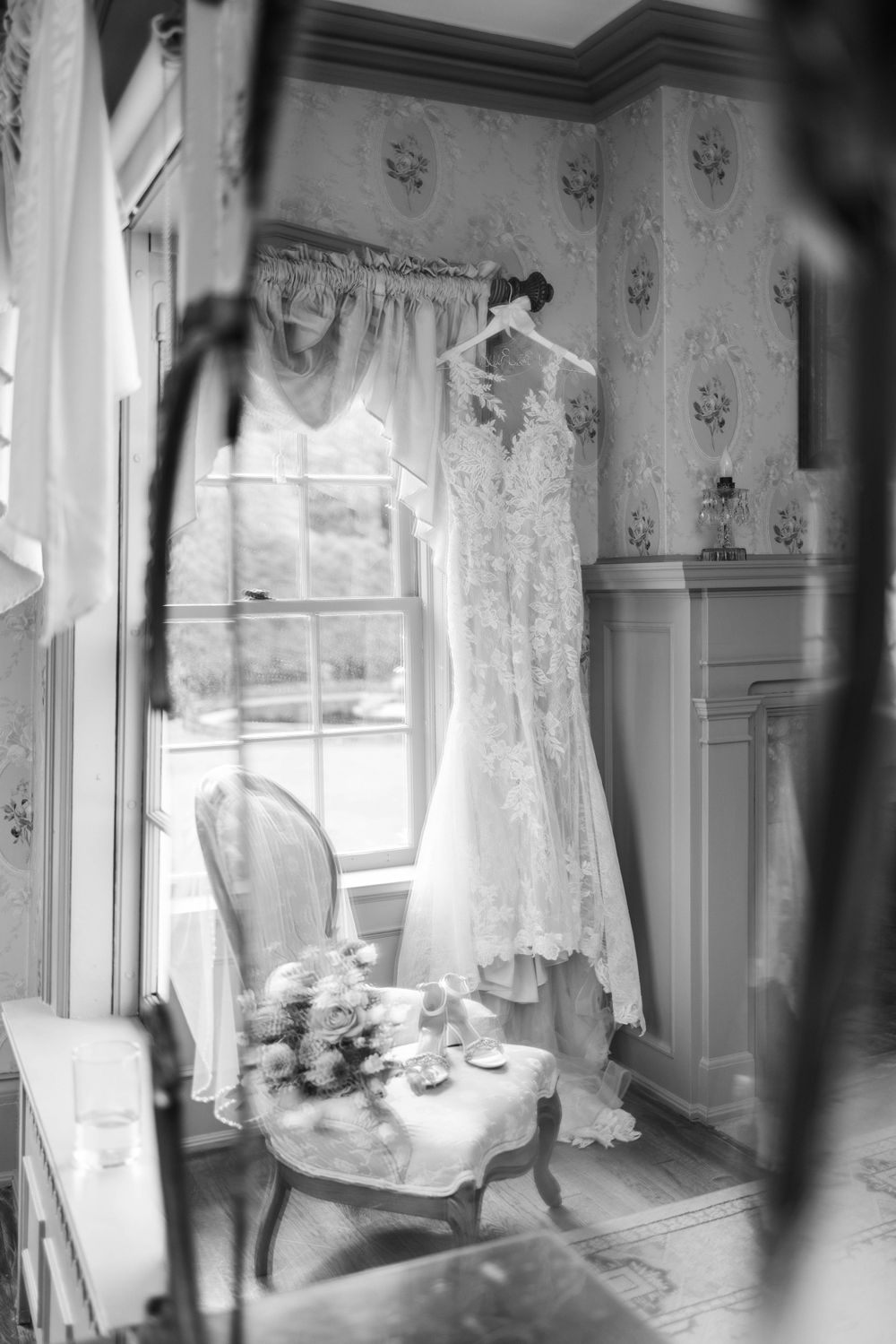 Black and white photo of wedding dress hanging by window with vintage chair and table setting.