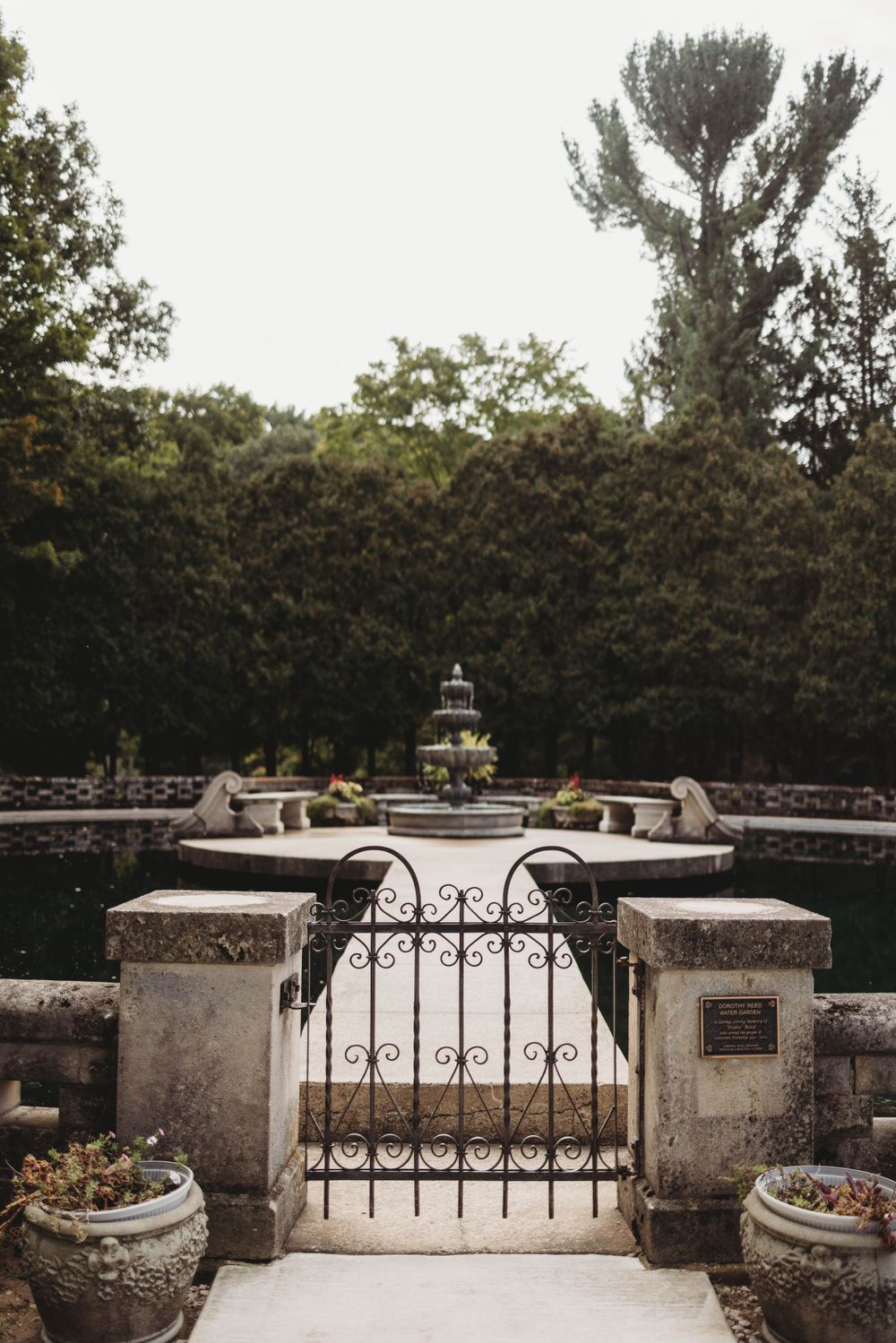 Ornate iron gate entrance leading to a garden fountain with stone walls and potted plants.