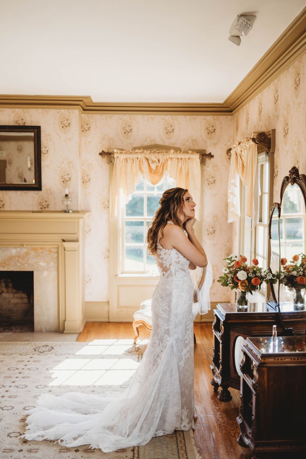 Bride in fitted lace gown poses by window in sunlit vintage room with floral wallpaper.