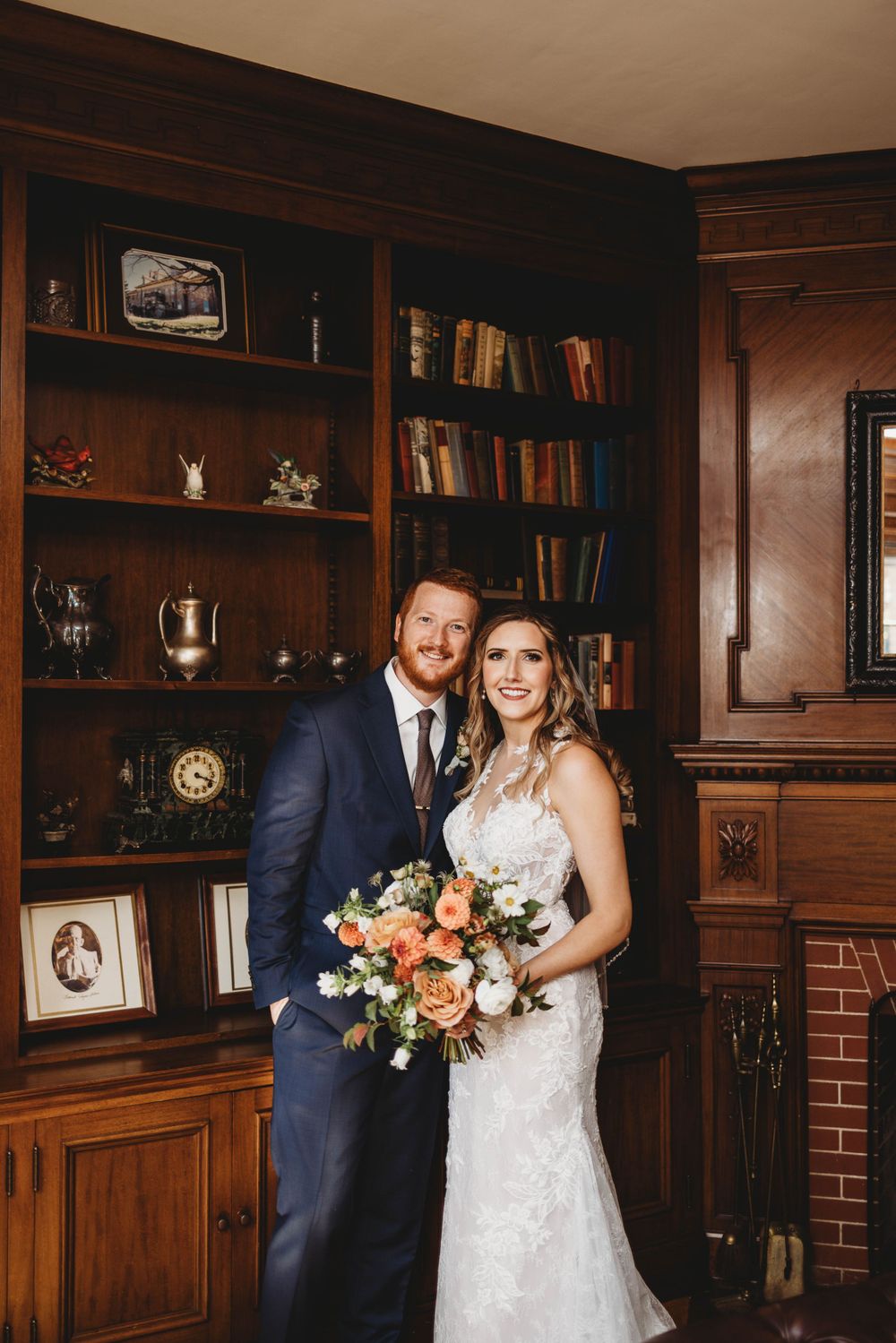 Newlyweds posing in vintage library with wooden bookshelves and brick fireplace.