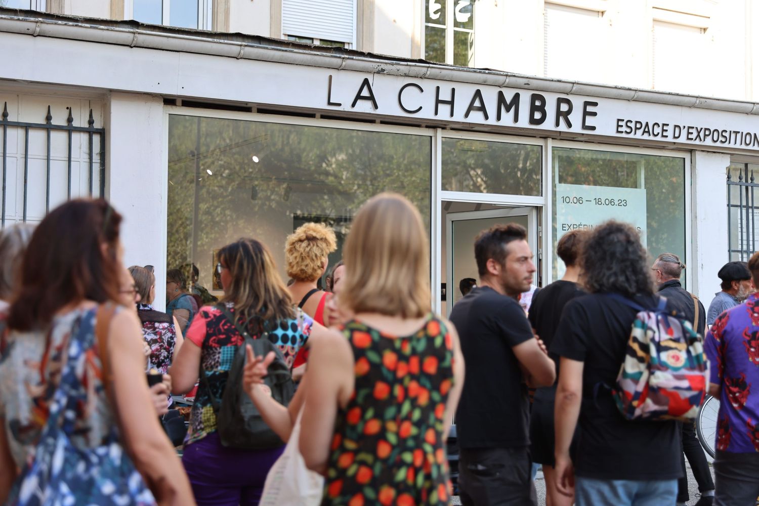 People gathering outside LA CHAMBRE building with crowd in summer attire.