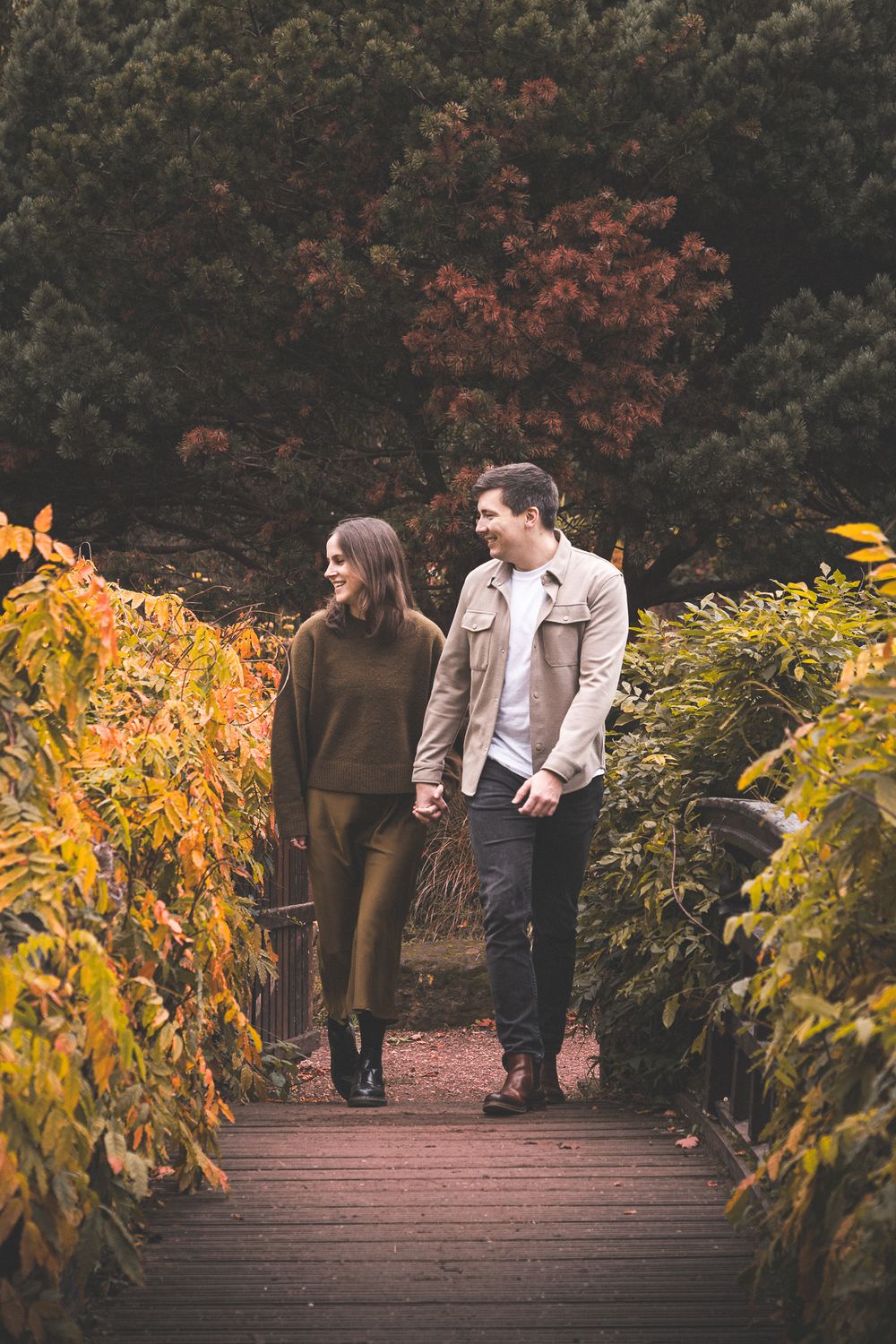 A couple walking and laughing together on a wooden bridge surrounded by fall foliage.