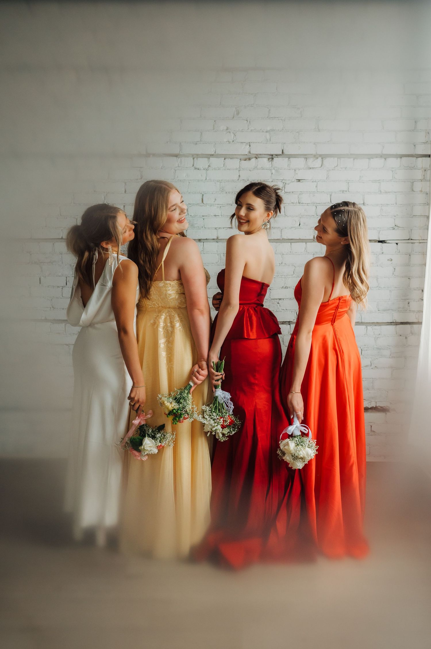 Four bridesmaids in elegant dresses of white, yellow, red and orange pose against white brick wall.
