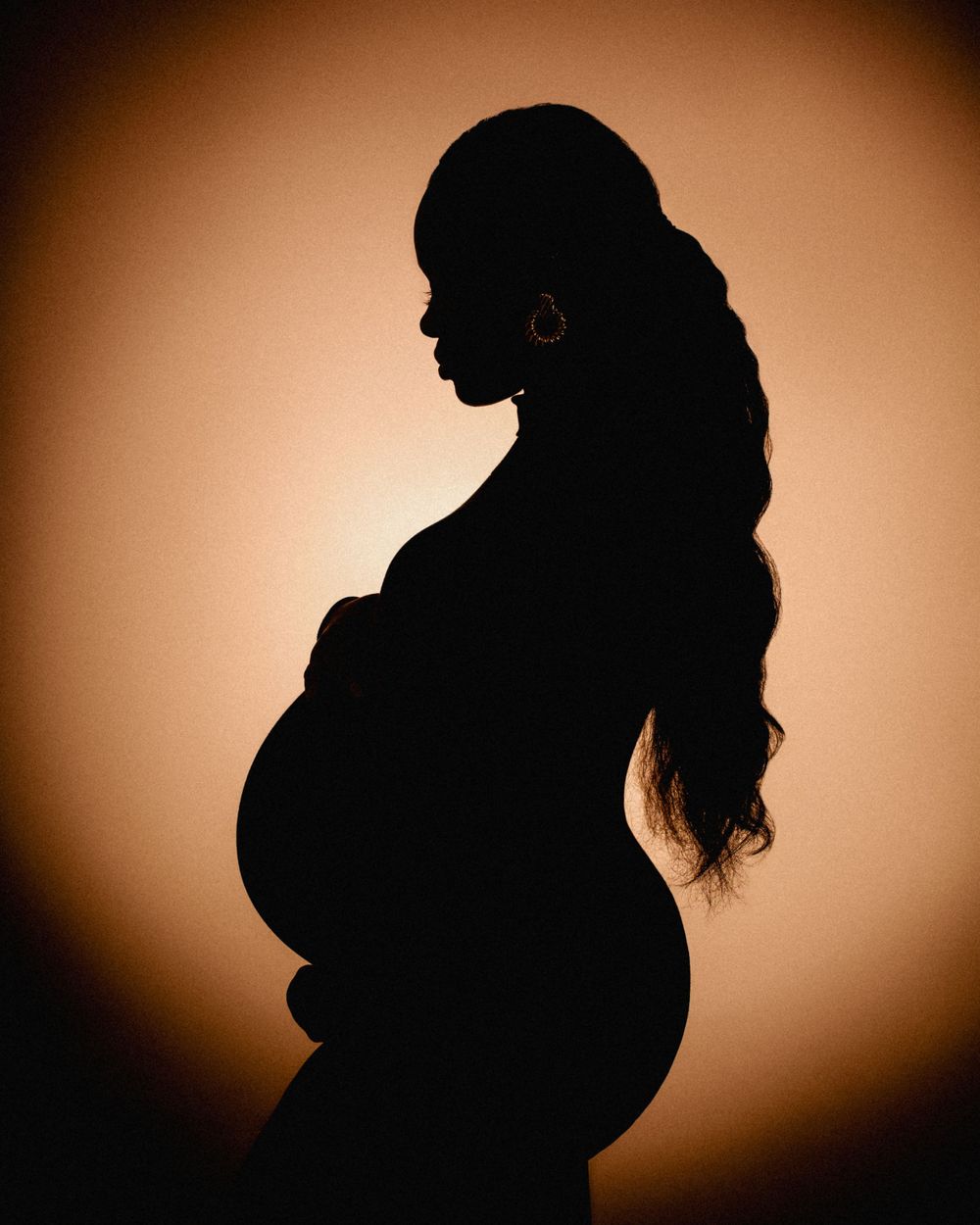 Silhouette profile of a pregnant figure with long flowing hair against a warm backlit background.