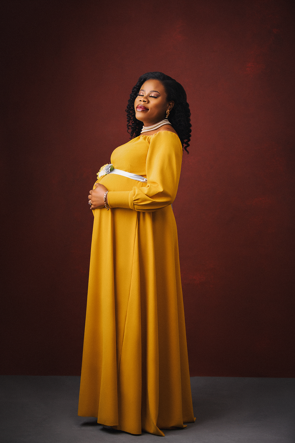 A maternity photo featuring a flowing yellow maxi dress against a dark red background.