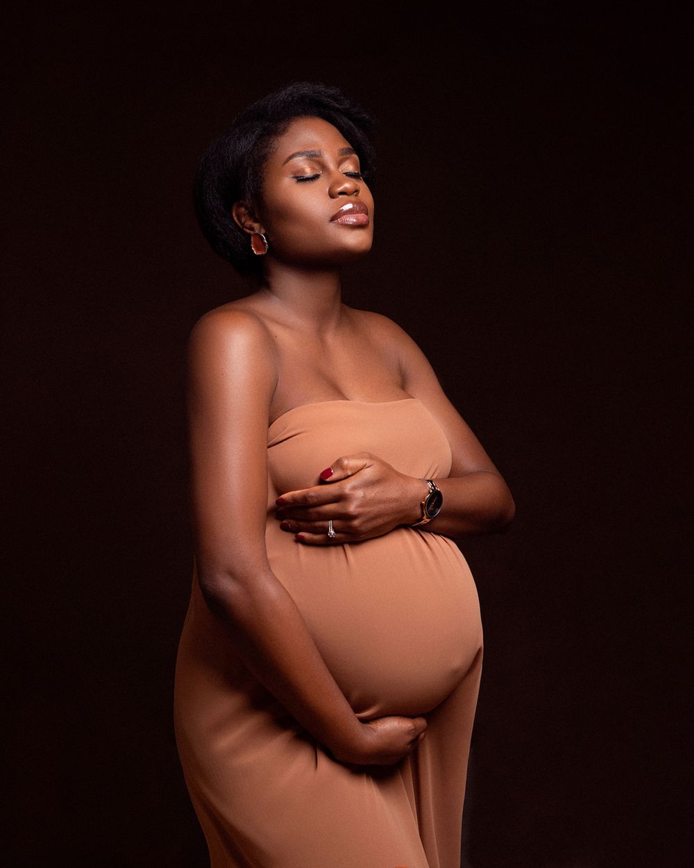 A pregnant woman in a brown studio portrait poses gracefully with closed eyes in warm lighting against a dark backdrop.
