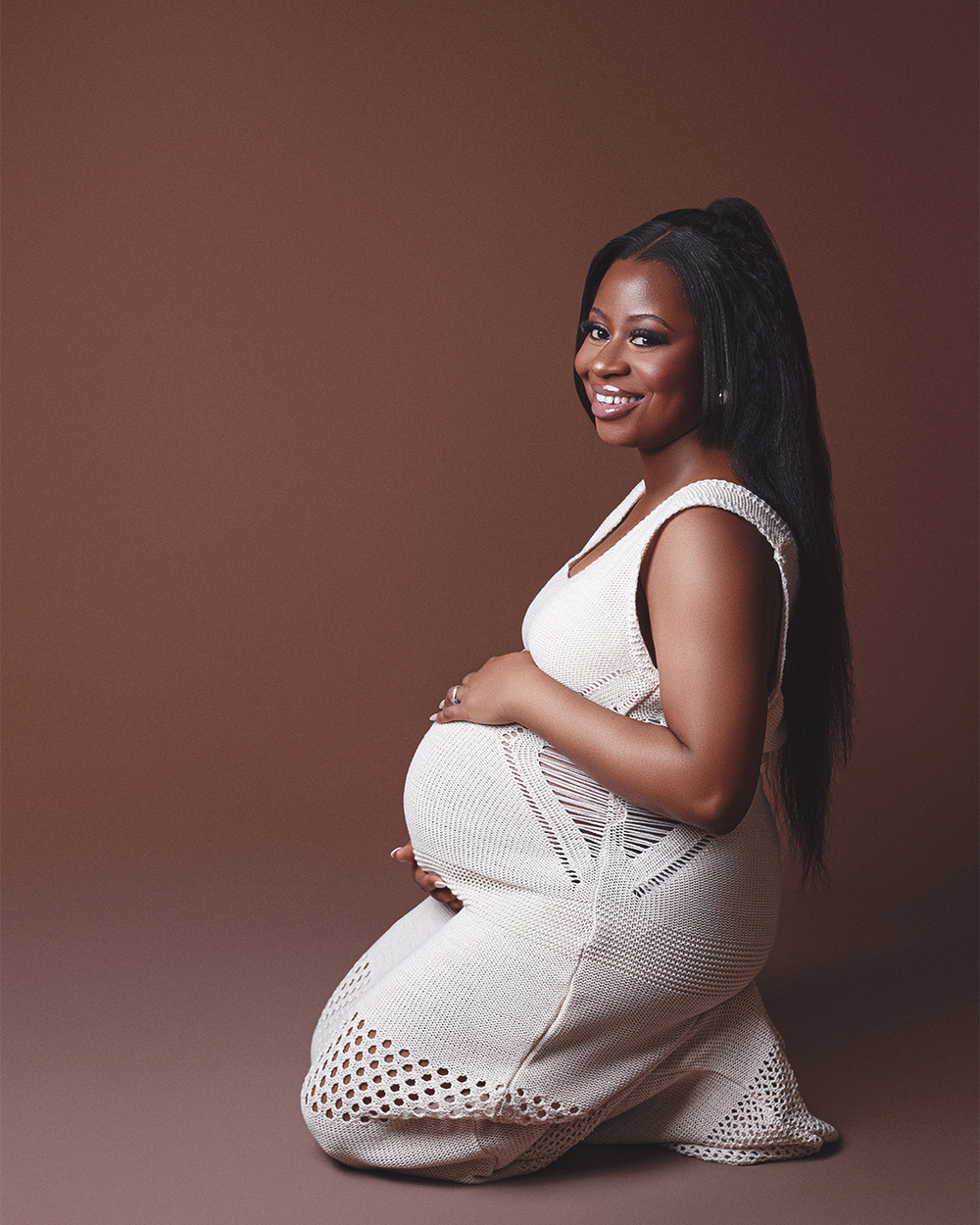 A maternity portrait in a white textured dress against a brown backdrop.