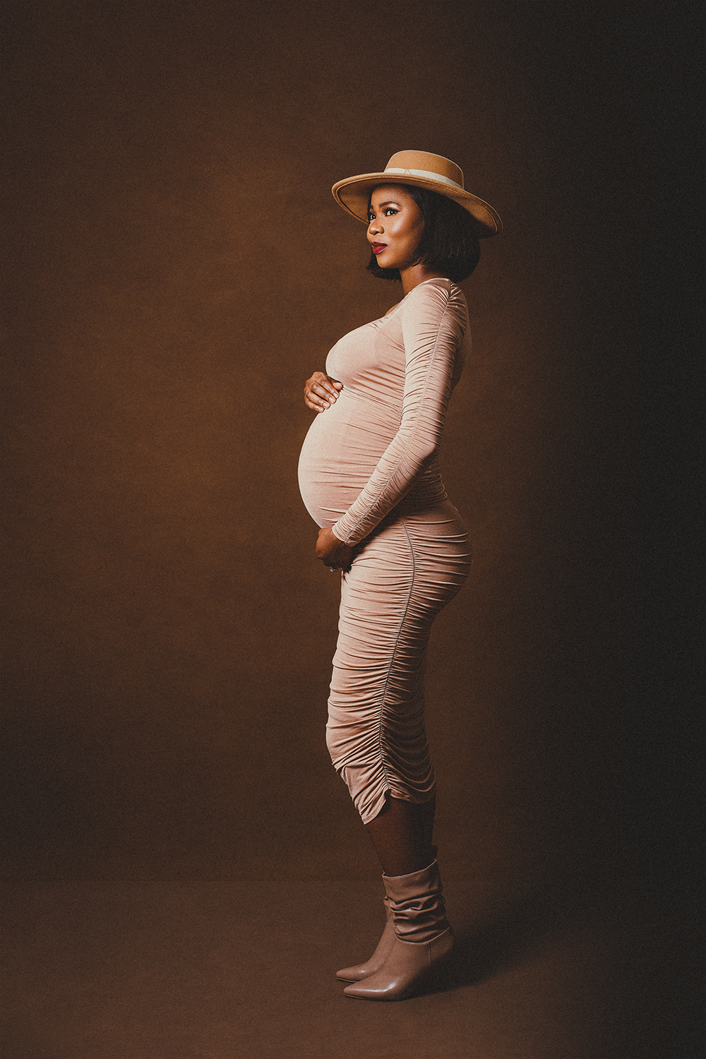 A maternity photo shows an expectant mother in a beige ruched dress and hat against a dark background.