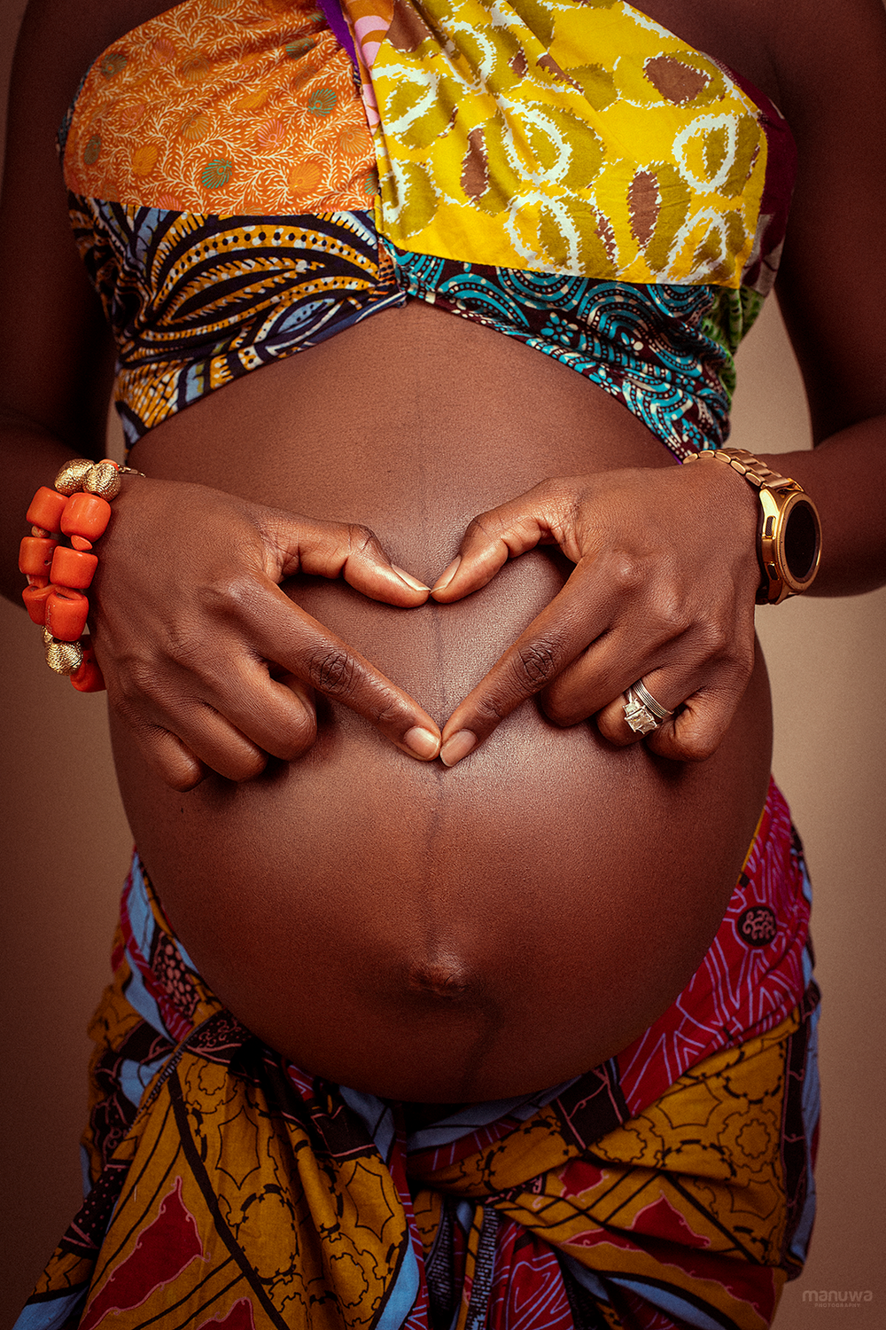 Close-up of pregnant belly with hands forming heart shape, wearing colorful African print fabric and jewelry.