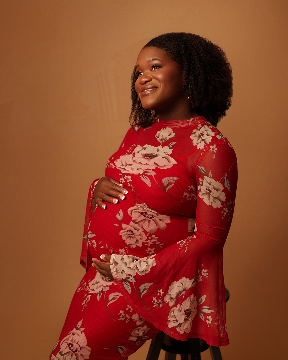 Person in a red floral dress poses against a warm brown background with natural curly hair.