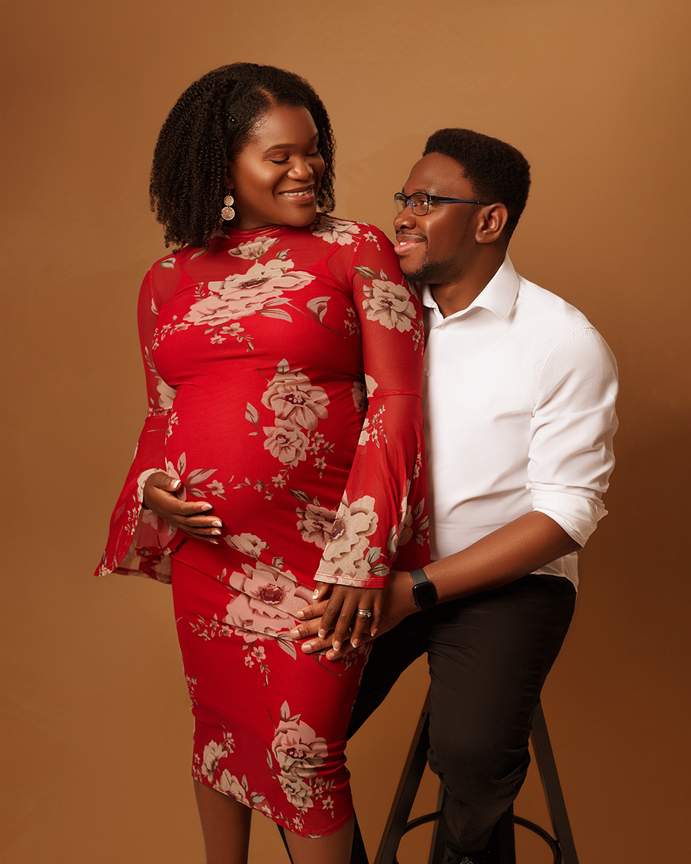 A couple poses in a studio against a brown backdrop wearing a red floral dress and white button-down shirt.