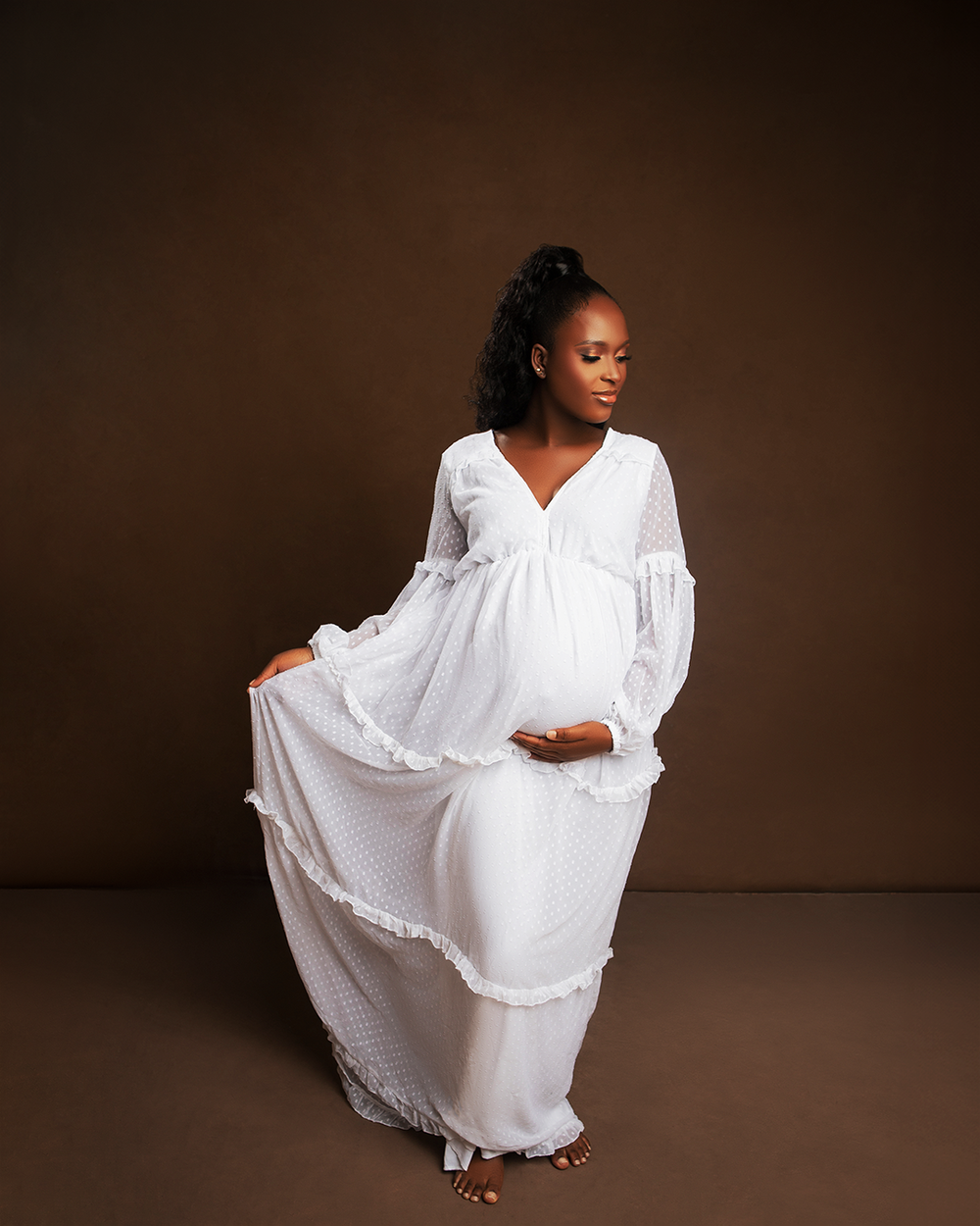 Elegant maternity portrait featuring a flowing white maxi dress with lace details against a dark brown background.