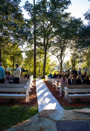Outdoor wedding ceremony setup with white benches and decorations under tall trees at sunset.