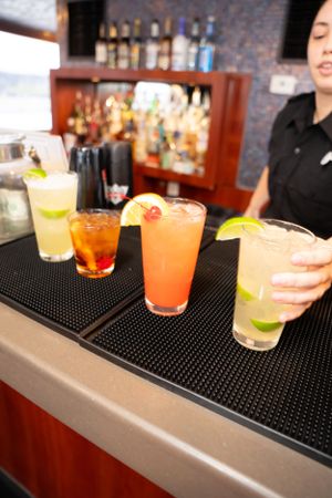 Colorful cocktails lined up on black rubber bar mat with citrus garnishes.