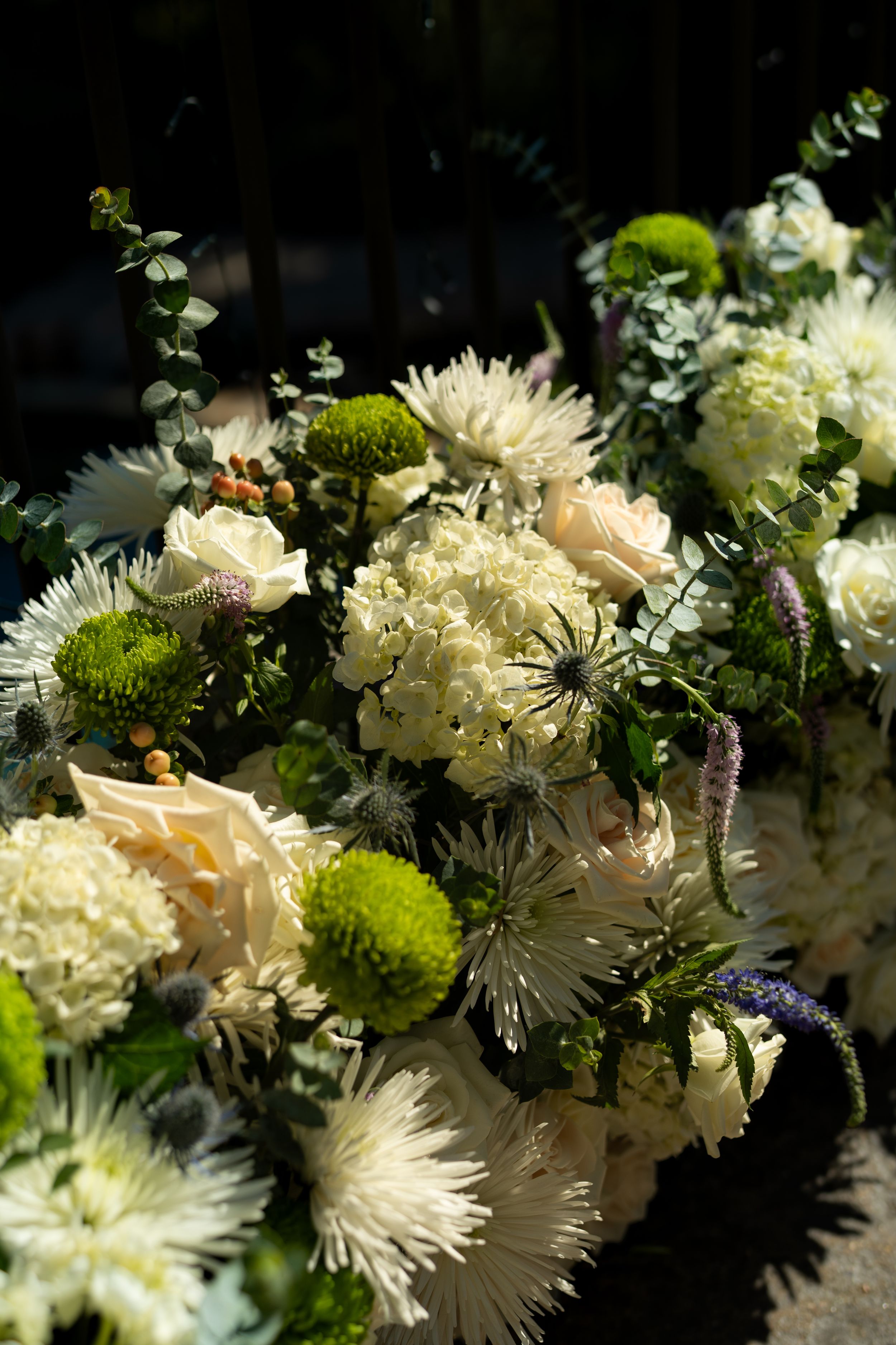 Delicate white and cream floral arrangement with spider mums, roses and green accents against dark background.