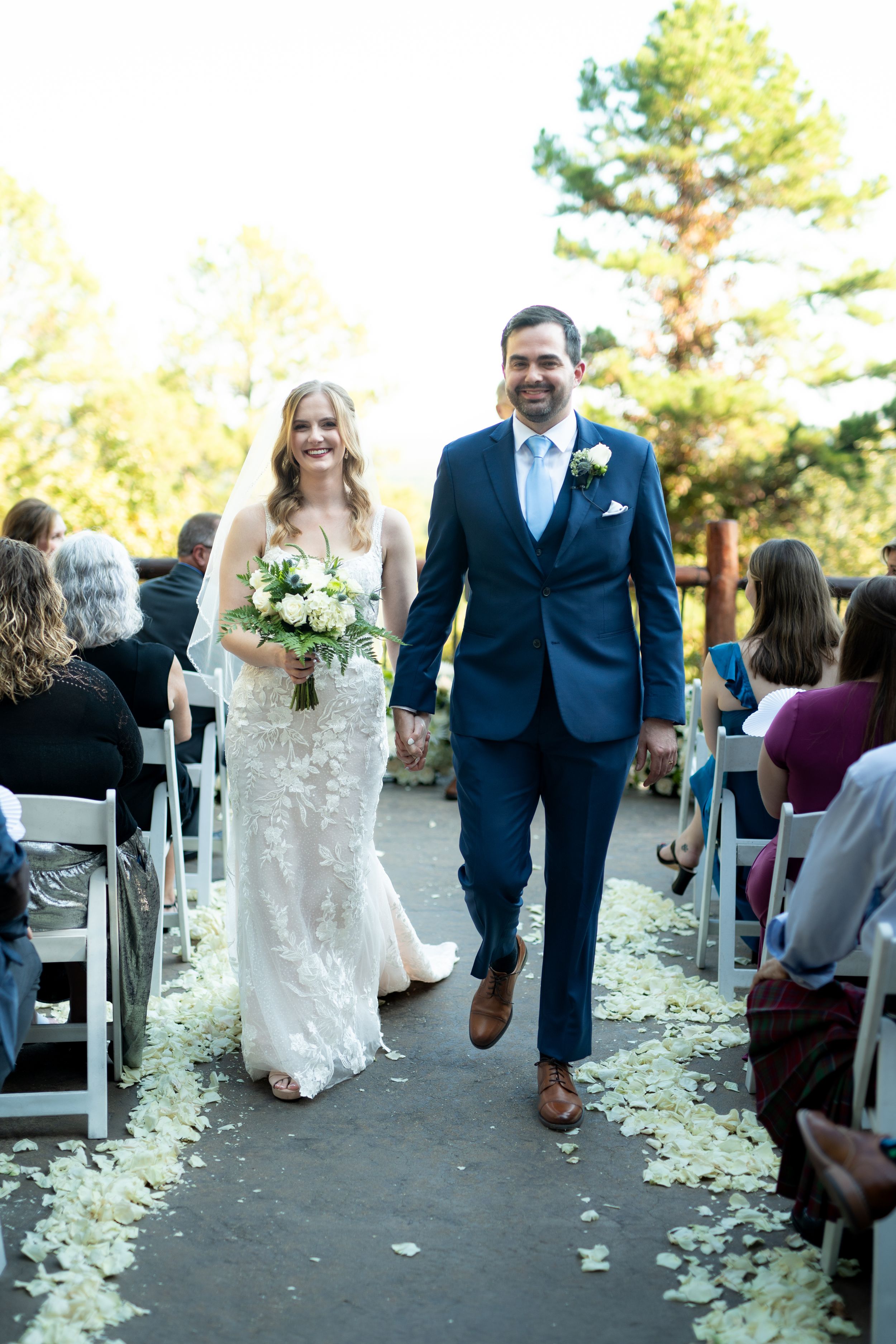 Newlyweds walking down the aisle lined with white flower petals and seated guests.