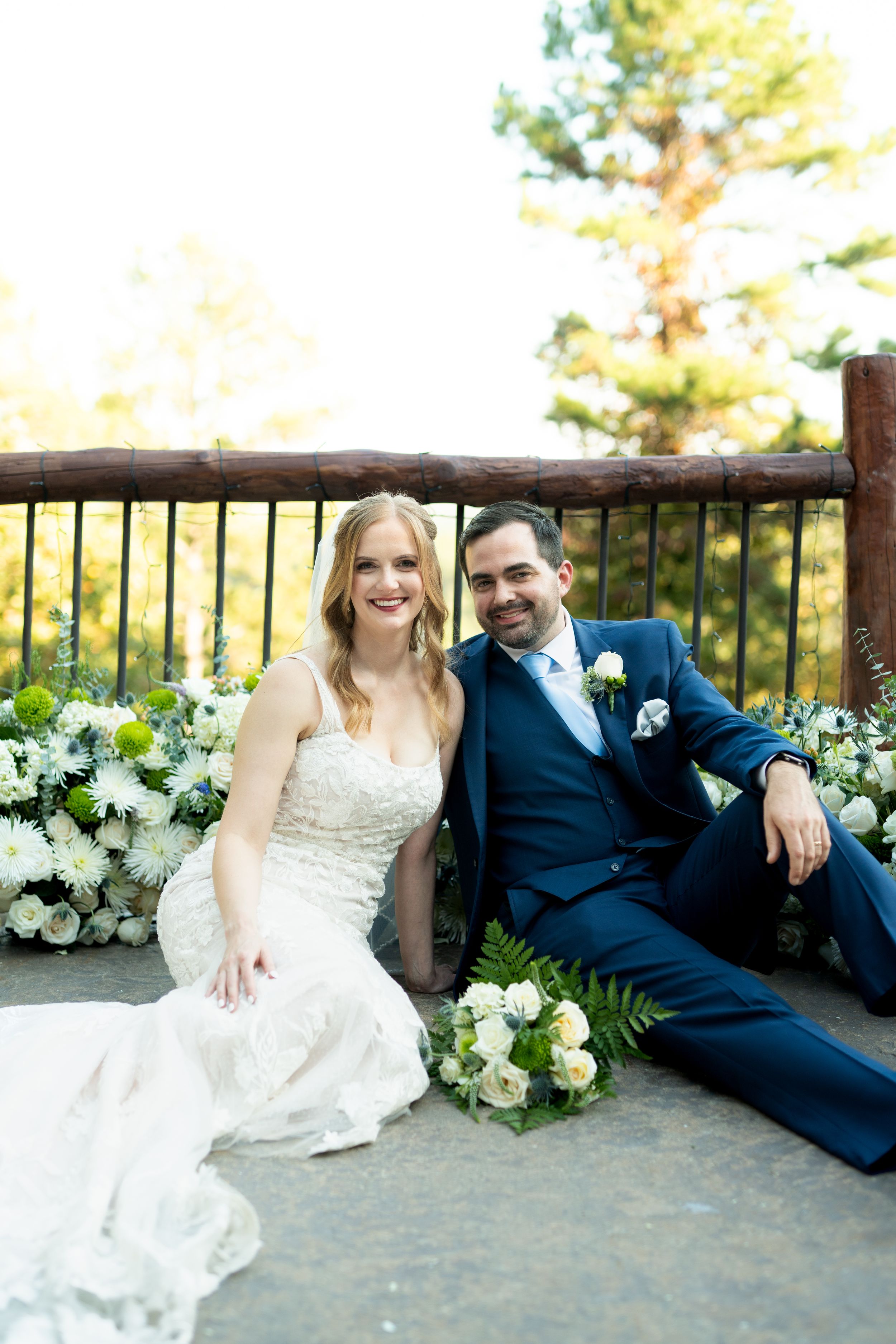 Newlyweds sitting together on rustic wooden bench with white floral arrangements.