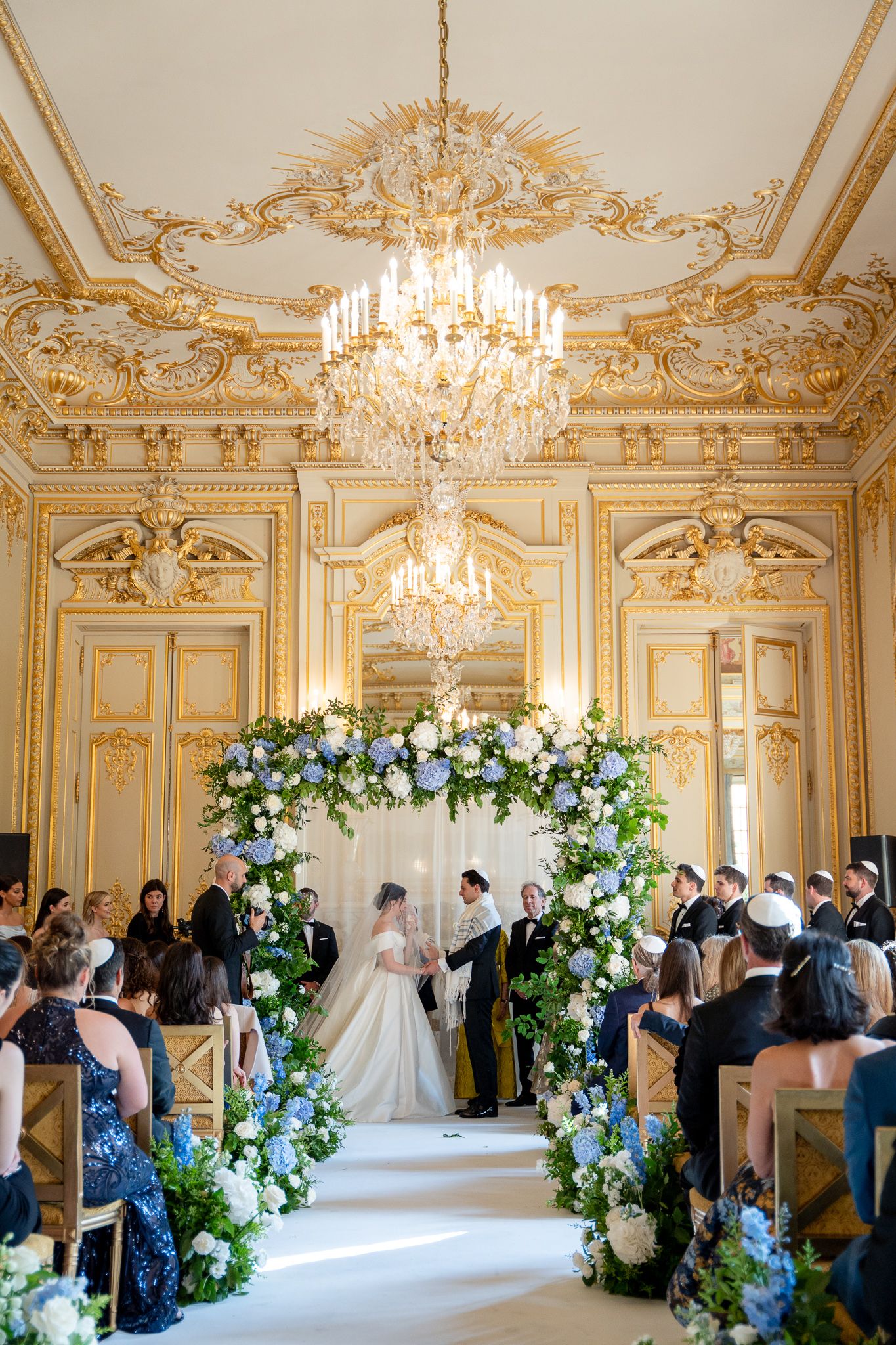 Elegant wedding ceremony in an ornate ballroom with crystal chandeliers, gold trim, and floral arches along the aisle.