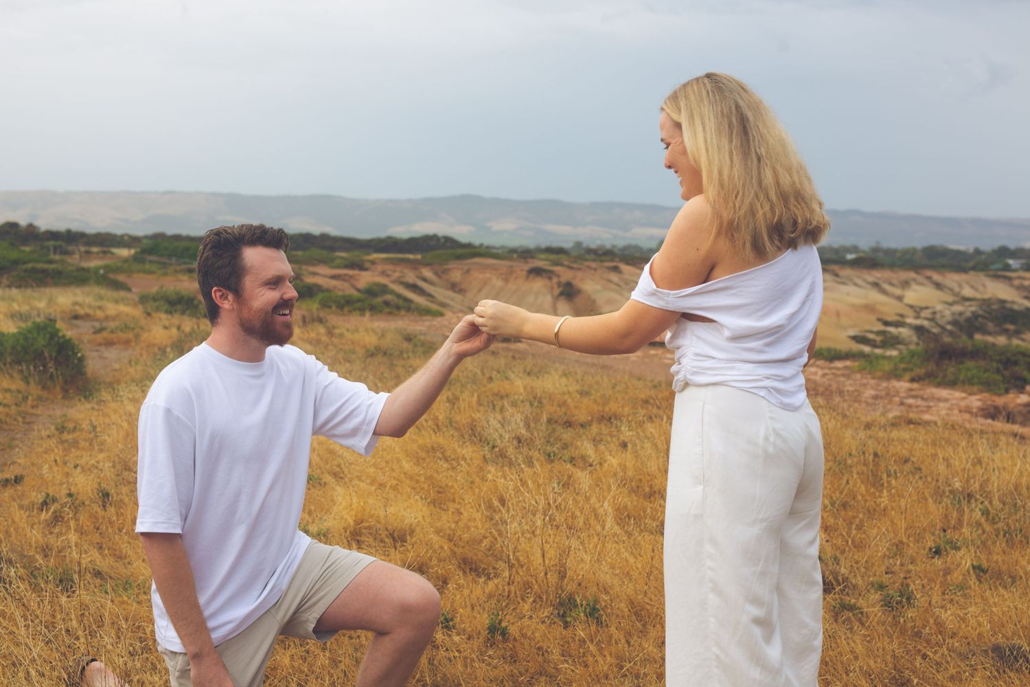 A romantic proposal scene unfolds on a coastal hillside with rolling hills in the background.