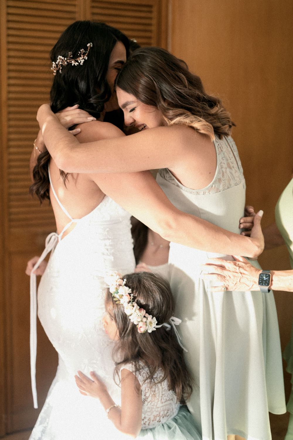 Three people in white dresses share an emotional embrace during a wedding celebration.