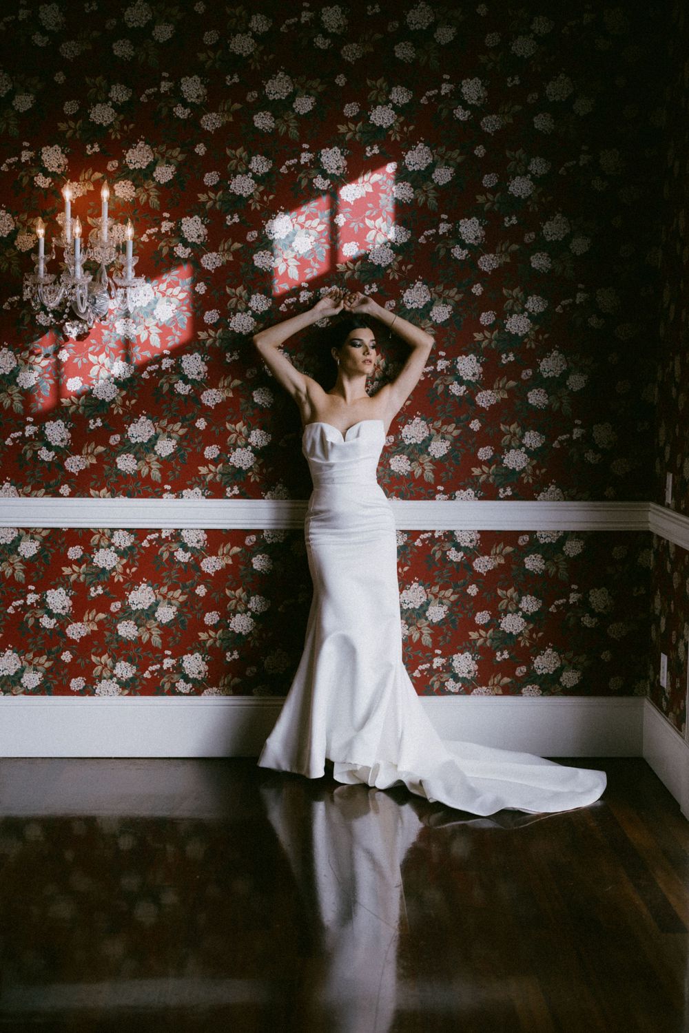 Dramatic bridal portrait against floral wallpaper with candles and fitted white gown.