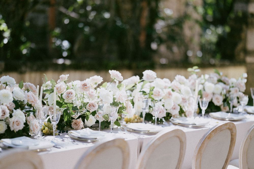 Elegant wedding reception table decorated with white floral centerpieces and cream chairs in an outdoor garden setting.