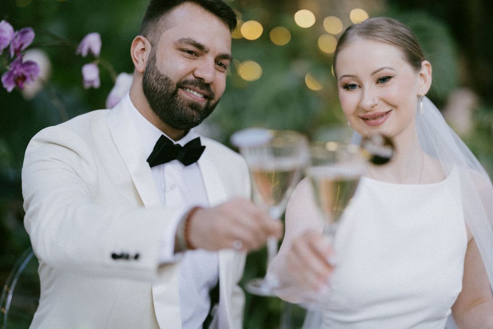 Newlyweds in white attire toasting with champagne glasses in garden setting.