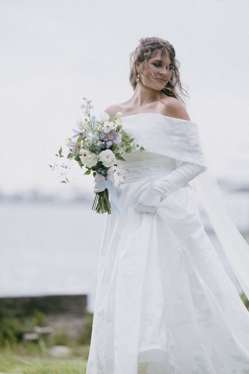 Ethereal bridal portrait with off-shoulder gown and delicate bouquet against misty background.