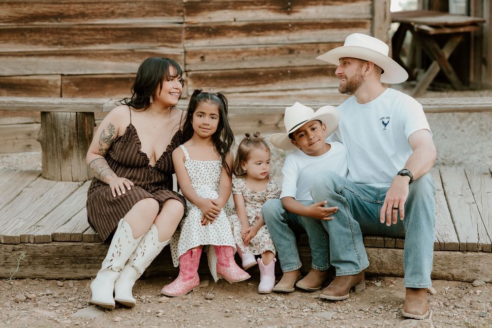 Family of five sitting on rustic cabin steps, parents in western attire with cowboy hats and boots, three young children smiling.