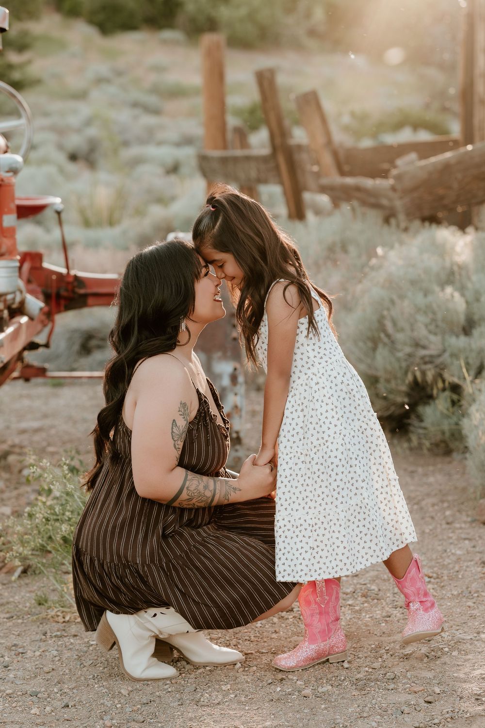 A mother crouches down to share a tender moment with her young daughter in pink cowboy boots outdoors.