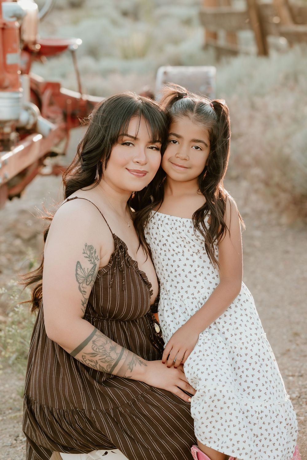 A woman in a brown dress and a young girl in a white floral dress pose together outdoors near a red tractor.