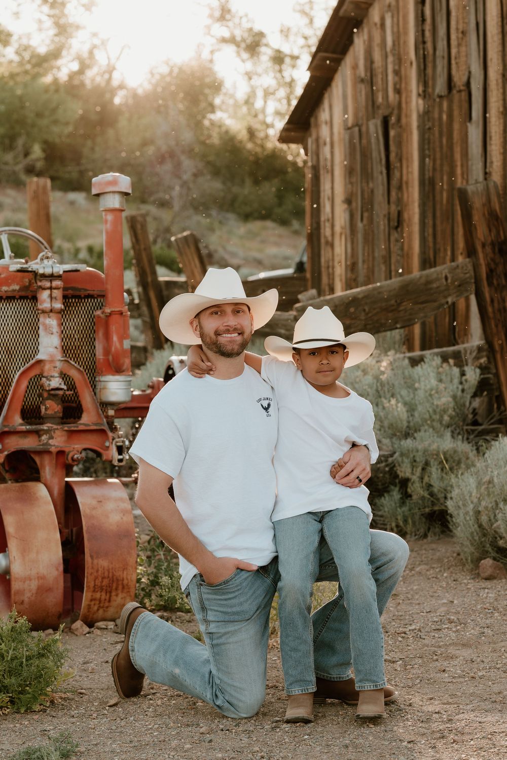 A man and boy in matching white tees, cowboy hats, and jeans pose by a vintage red tractor near a rustic barn.