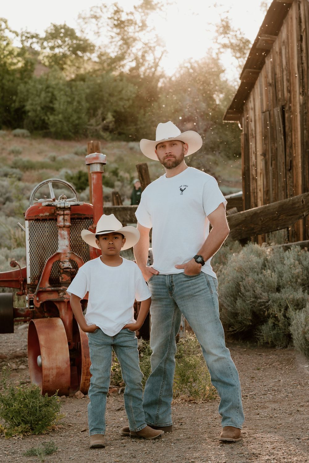 A man and boy in matching white shirts, jeans, and cowboy hats pose near a vintage red tractor and rustic barn.