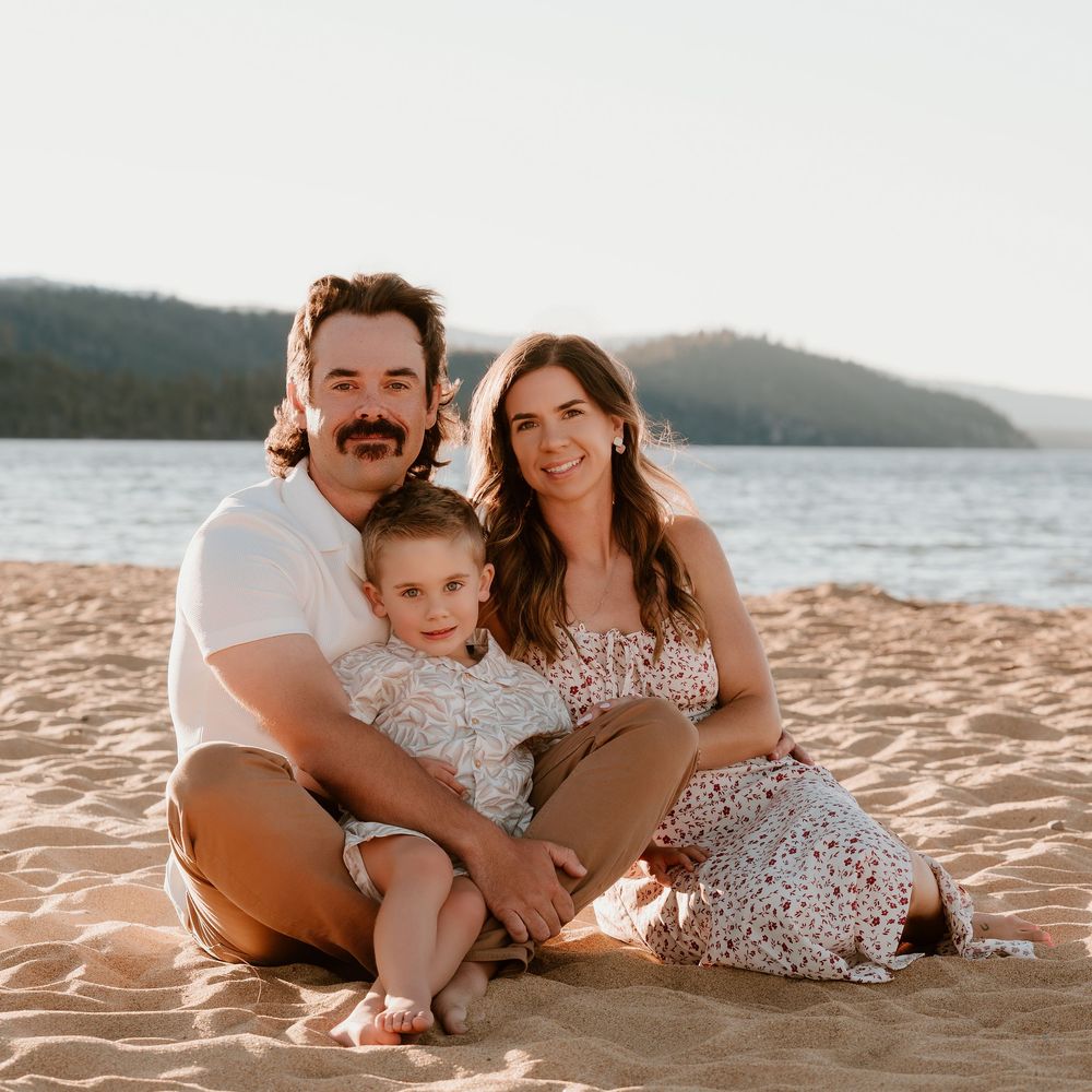 A happy family of three sits together on a sandy beach at golden hour with a lake and hills in the background.