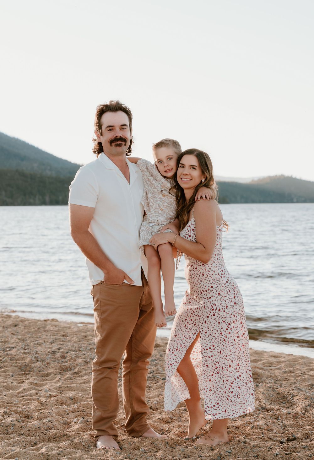 Family of three smiling on a sandy beach at sunset, with a lake and mountains in the background.