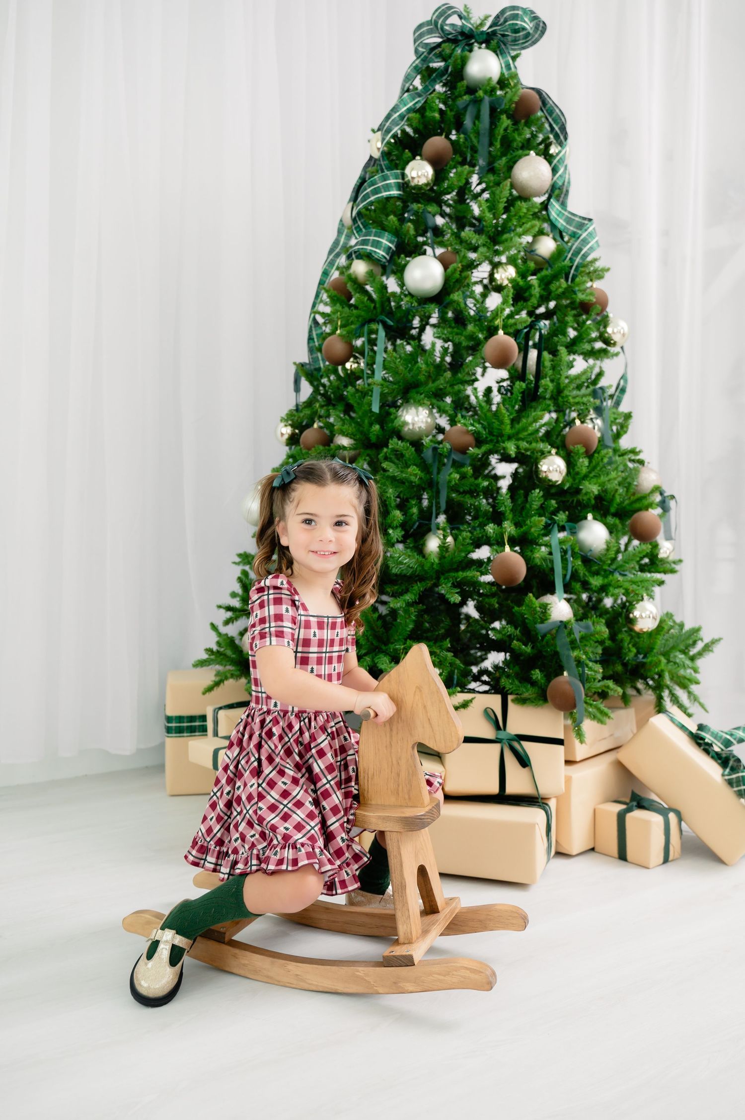 Beautifully decorated Christmas tree with presents underneath stands behind a wooden rocking horse toy in a bright room.