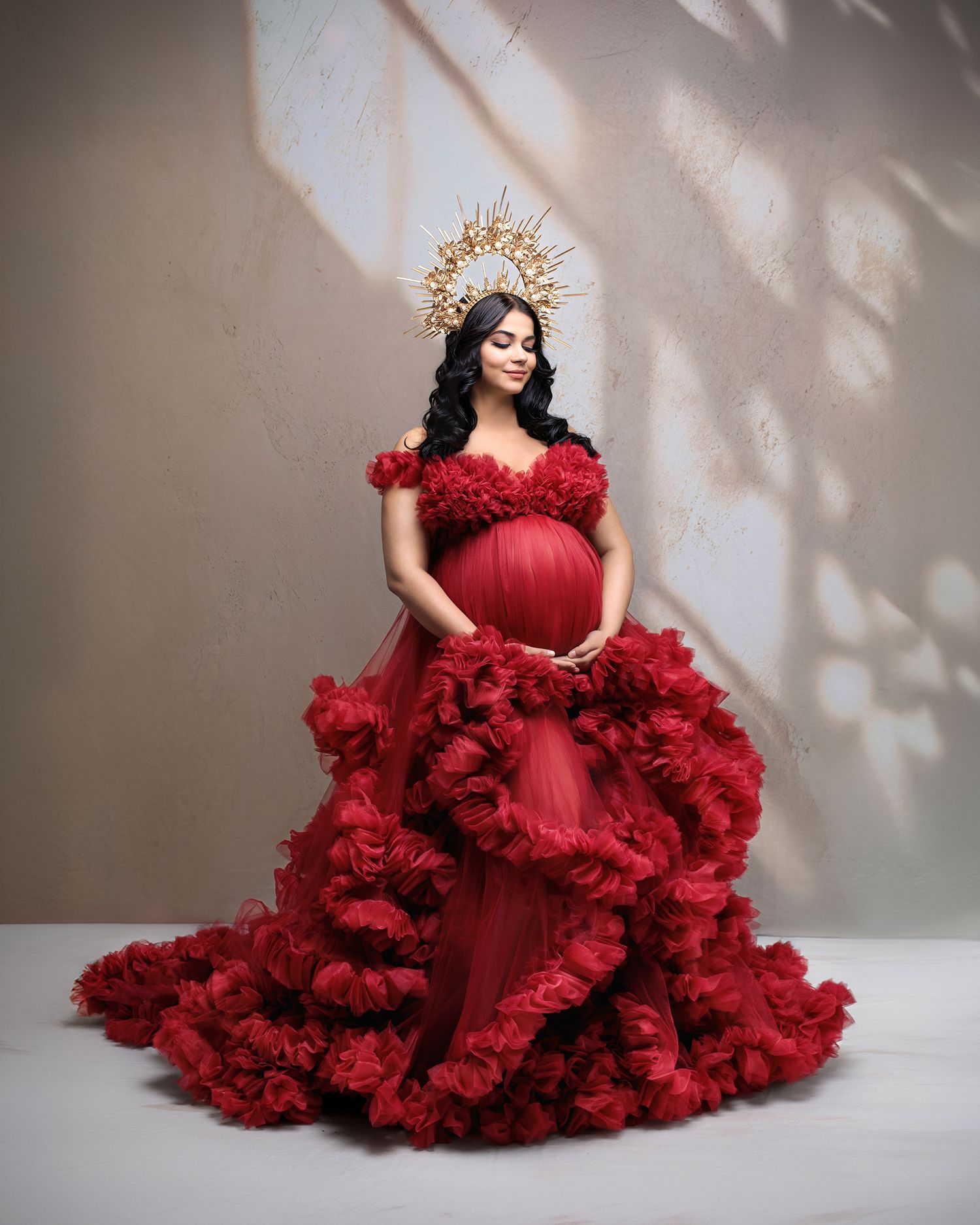 A greek goddess inspired maternity portrait the woman wearing a long flowing white gown against a honey lit shadow wall.