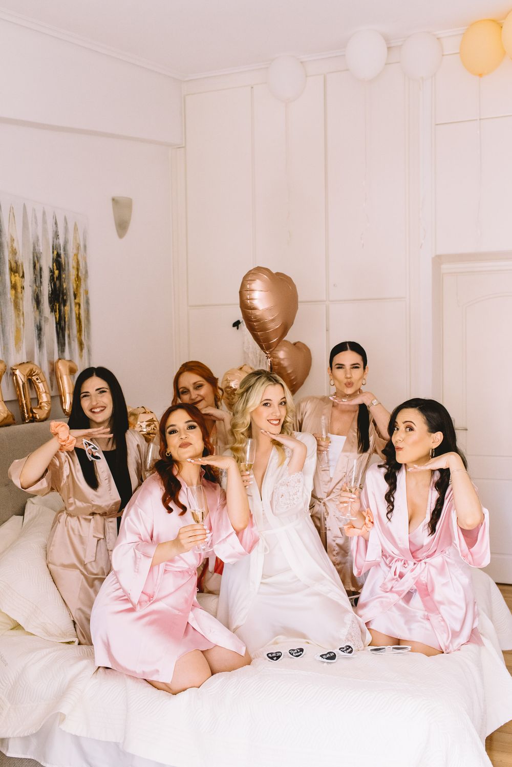 A group of friends in matching pink satin robes celebrate at a cheerful slumber party gathering with balloons.