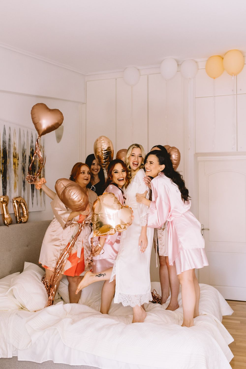 Group in pink robes celebrating on a white bed with gold and rose gold balloons decorating the festive space.