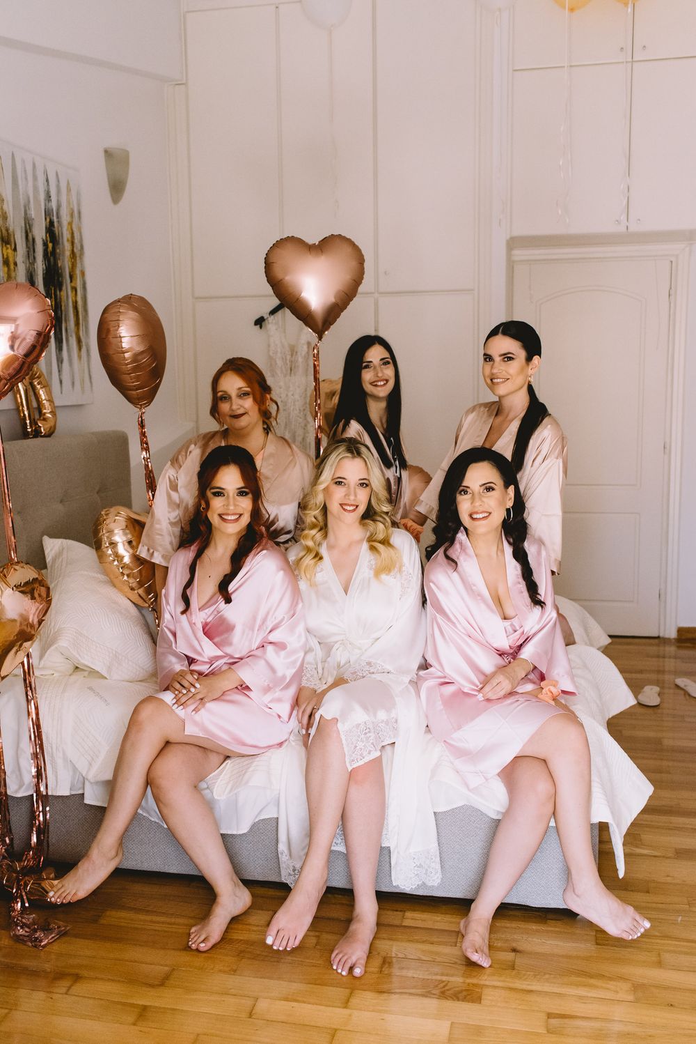 A group in matching pink satin robes pose together on a hardwood floor during a wedding day celebration.