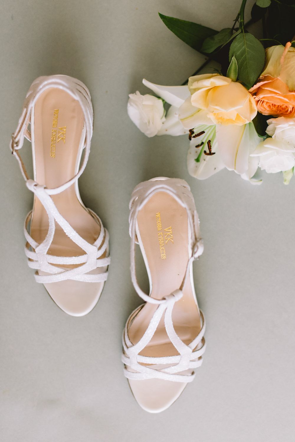 Elegant white strappy sandals with crossed design displayed next to peach and white roses on a light gray background.