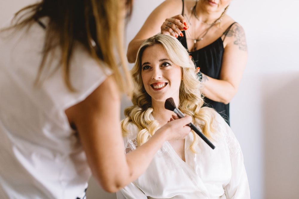 Stylist works on bride's hair during wedding preparations while she sits in white robe with makeup brush in hand.