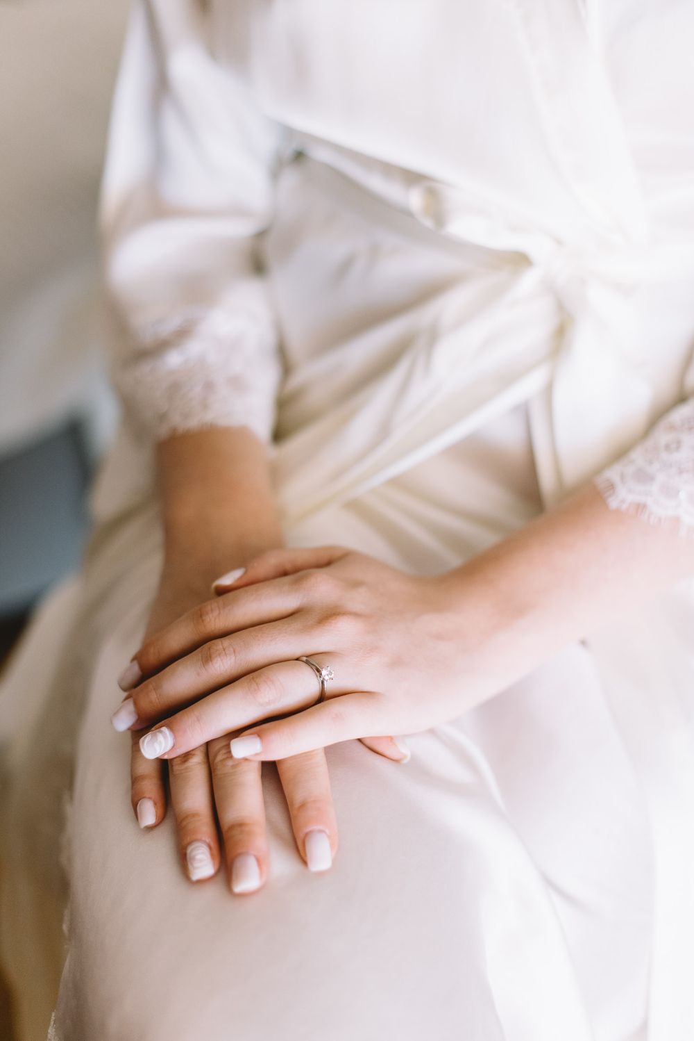 Close up of hands wearing a wedding ring against white satin and lace dress fabric with French manicured nails.