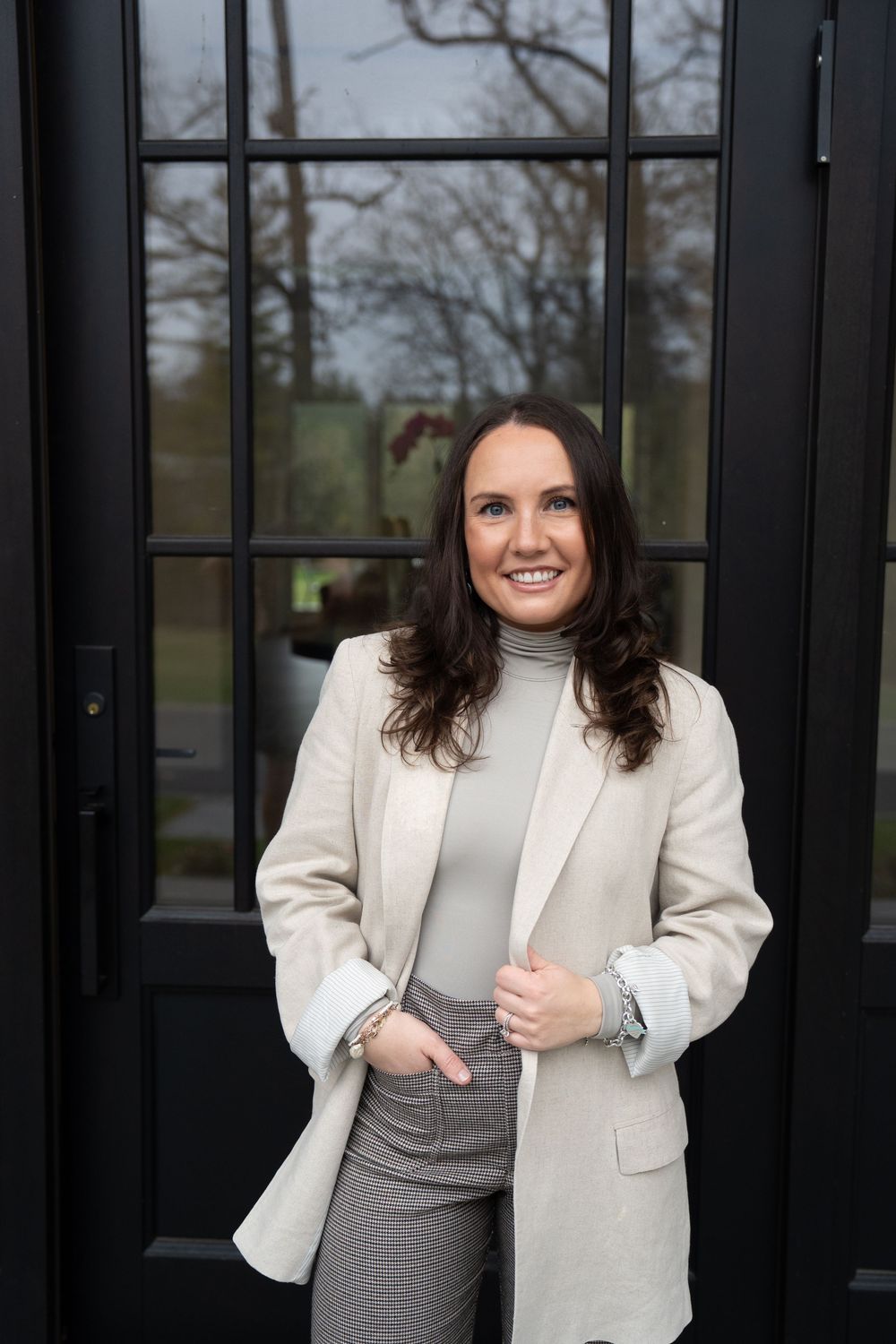 Professional wearing a white blazer and gray dress stands confidently against dark doors.