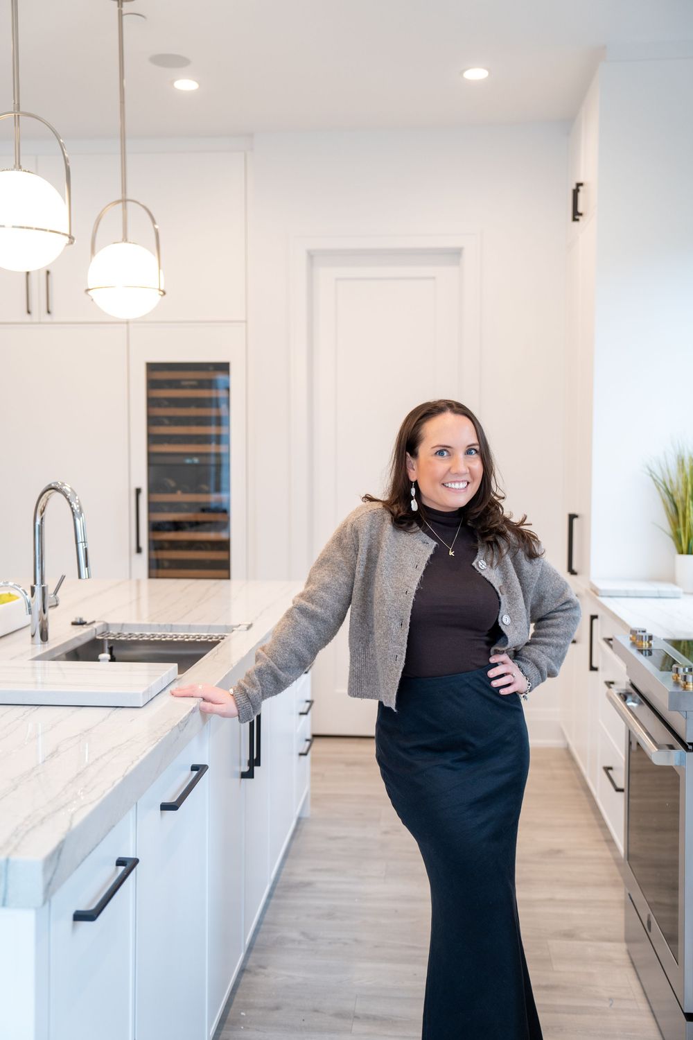 A person wearing a black dress and grey blazer poses in a modern white kitchen with light wood flooring.