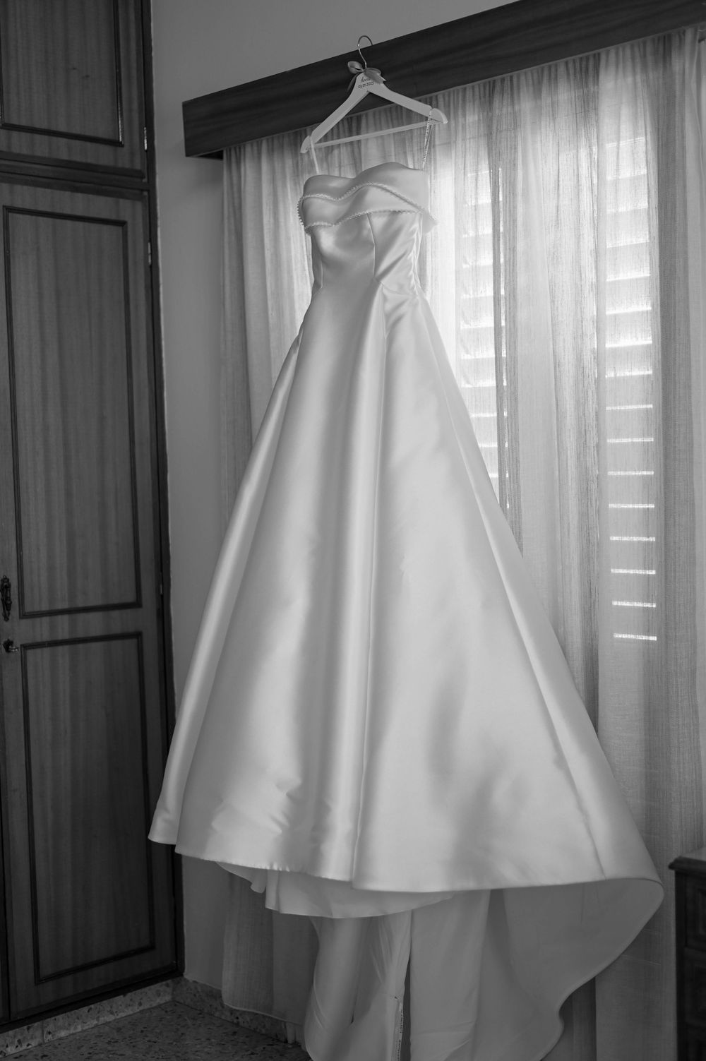 A classic white wedding dress with sweetheart neckline hangs elegantly by a window in black and white photography.