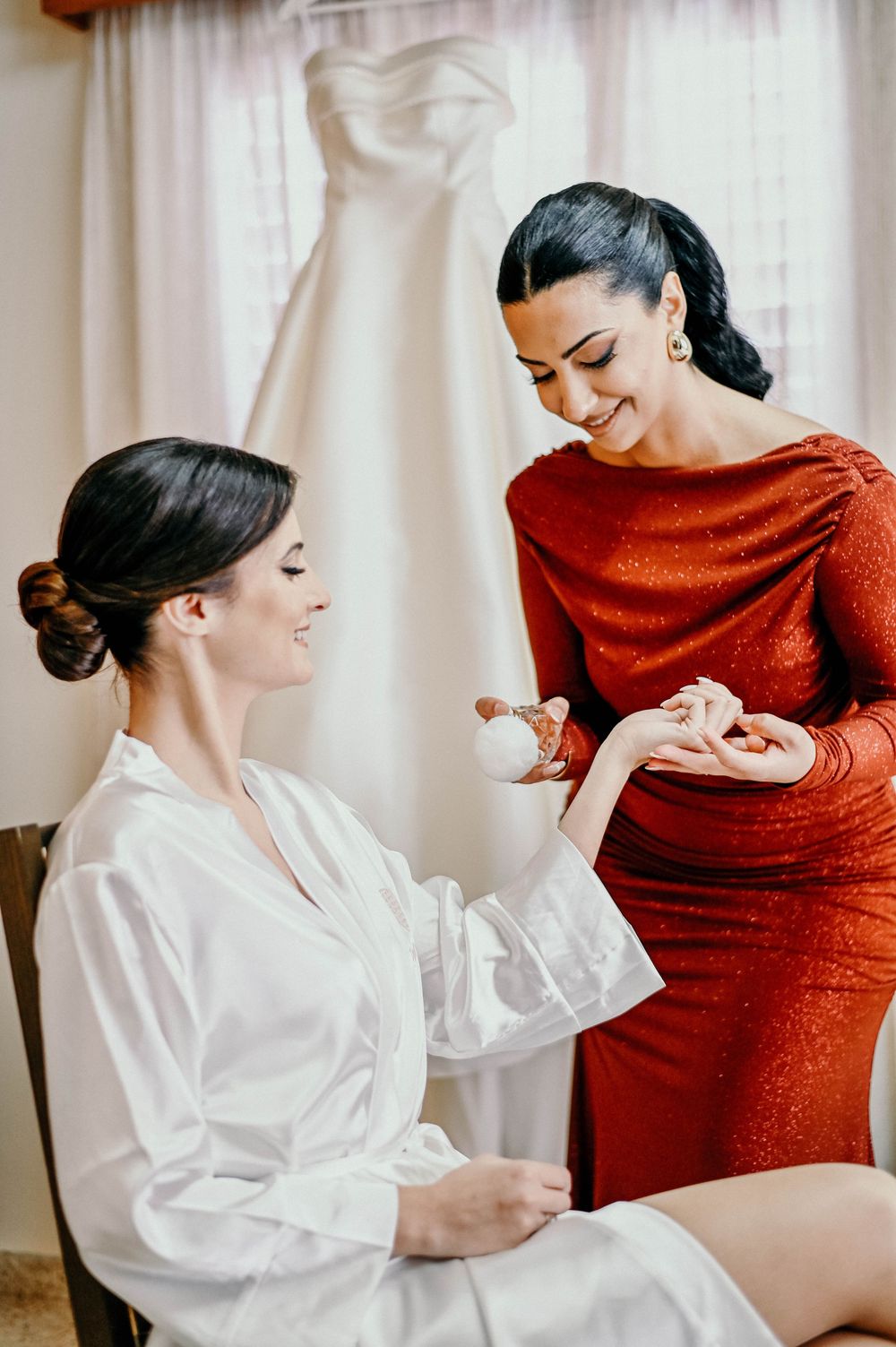 Two women share a joyful moment while getting ready, with a white dress hanging in background.