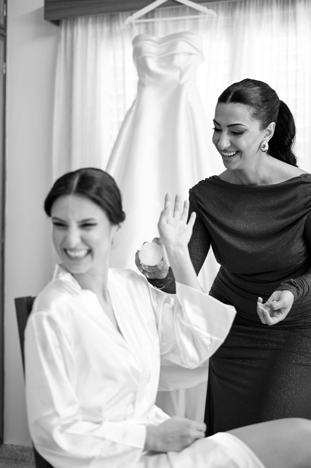 A bride and her bridesmaids share a joyful candid moment while getting ready in white robes and black dresses.