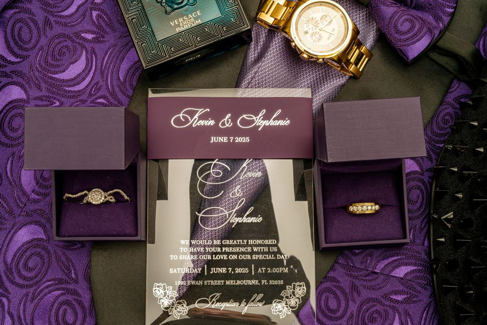 Wedding rings and invitation displayed with gold watch on purple textured background.